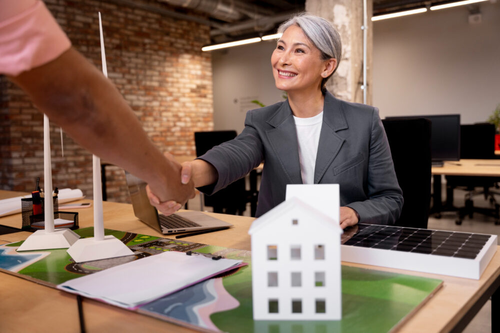 A smiling professional woman in a grey blazer shakes hands with another person across a desk with architectural models and documents, symbolizing increased sales and business success achieved through hiring a real estate virtual assistant.